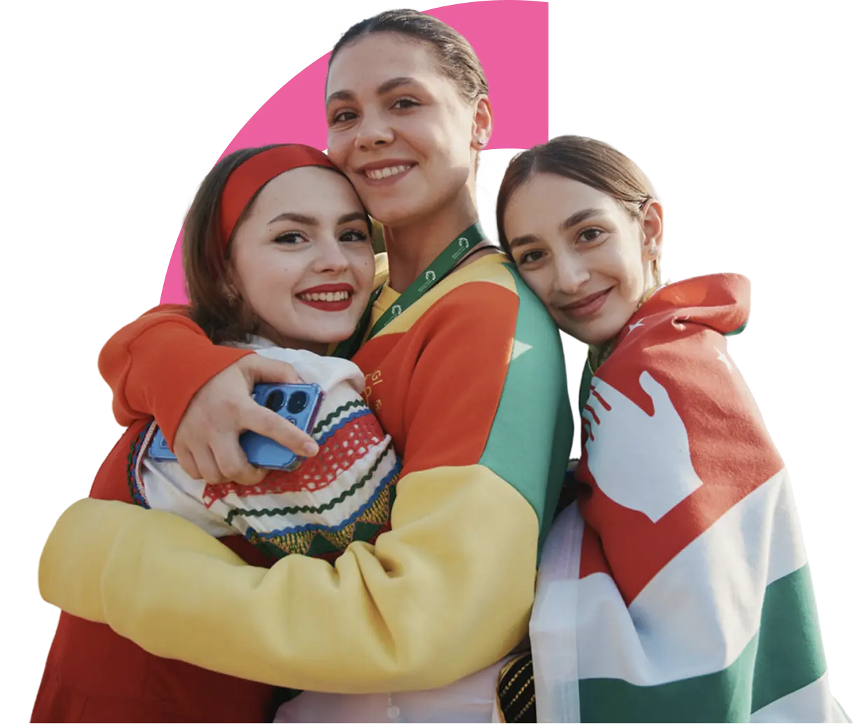 Three smiling young women hugging, one in traditional clothes, one in a colorful jacket, and one draped in a red, white, and green flag.
