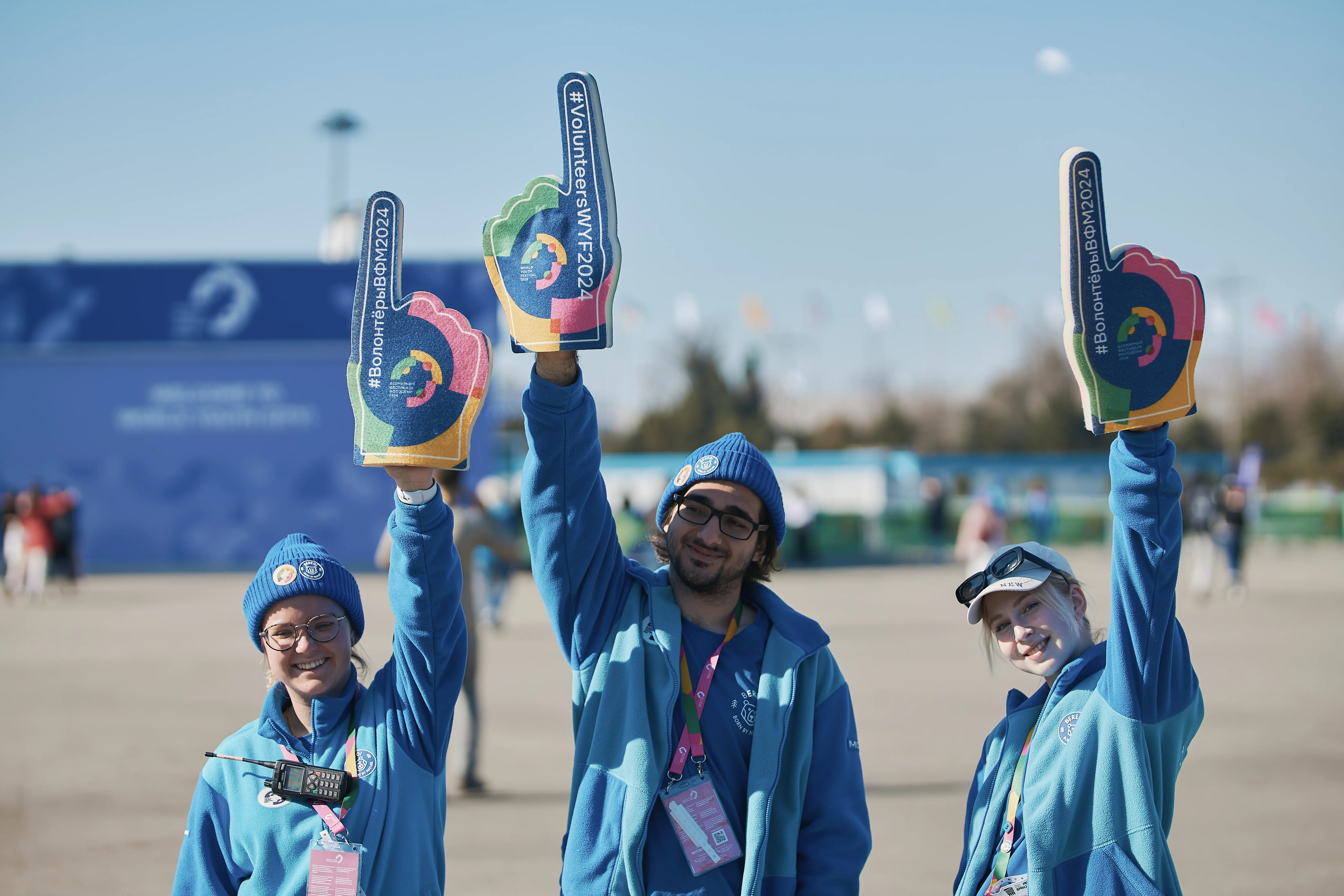 Улыбающаяся волонтёрка в оранжевой форме WYF держит большую пенную перчатку с надписью на русском языке. На фоне видны фиолетовые флаги мероприятия.