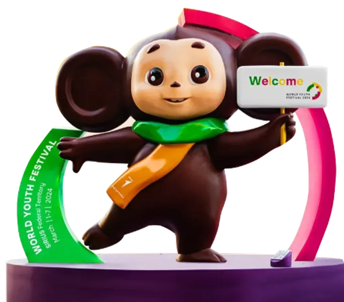 Чебурашка — знаменитый российский мультяшный персонаж с большими круглыми ушами — дружелюбно улыбается, держит табличку «Welcome» и цветные элементы оформления Всемирного молодёжного фестиваля.