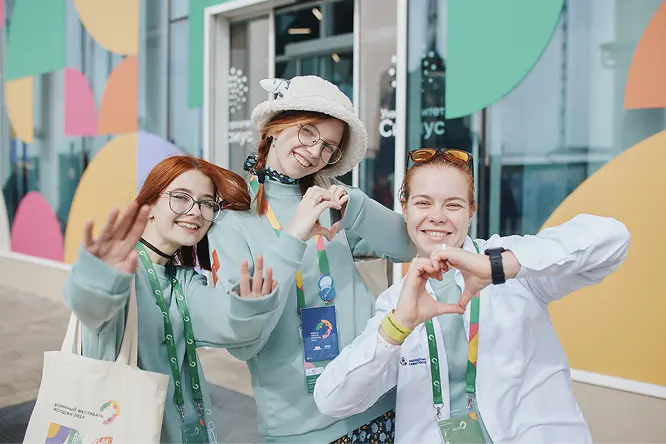 Три улыбающиеся молодые девушки на улице показывают жесты сердечек руками. Они носят яркие бейджи и сумки, позируют перед зданием с цветными геометрическими элементами.