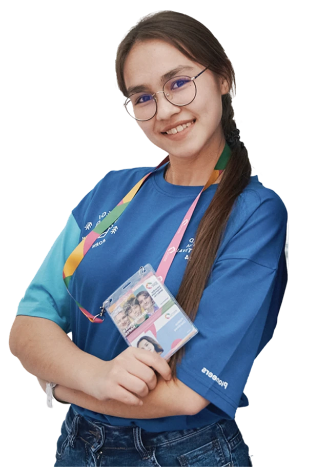 Молодая женщина с длинными заплетёнными волосами и круглыми очками, тепло улыбается в камеру, одета в яркую синюю волонтёрскую футболку с радужными полосами и держит бейдж на розовом шнурке.