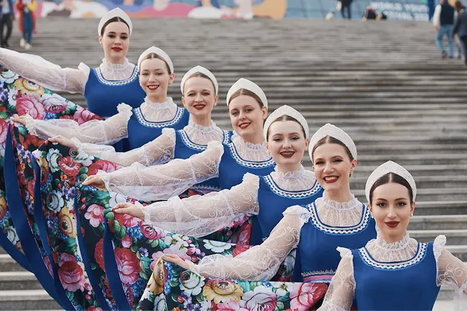 Группа танцовщиц в традиционных синих костюмах с цветными юбками стоит в ряд на широких ступенях, улыбаясь в камеру.